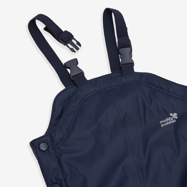 A close-up of Muddy Puddles' navy Puddleflex dungarees. The waterproof, fleece-lined trousers feature adjustable straps and reflective logo.